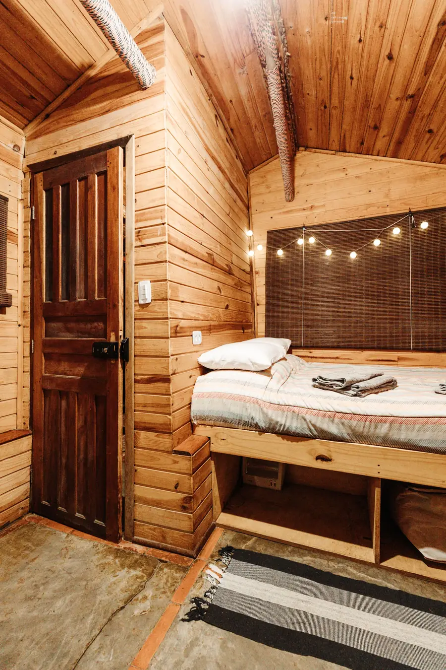 Quarto pequeno todo em madeira, com cama e cordão de luzes